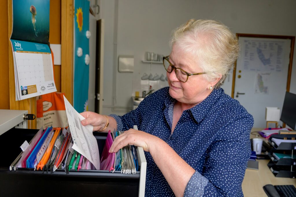 An older woman with glasses reviews files in a filing cabinet.
