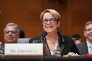 Karen Friedman sits at a table in the Senate HELP Committee hearing room. She has blond hair and blue eyeglasses. She wears a black blazer.